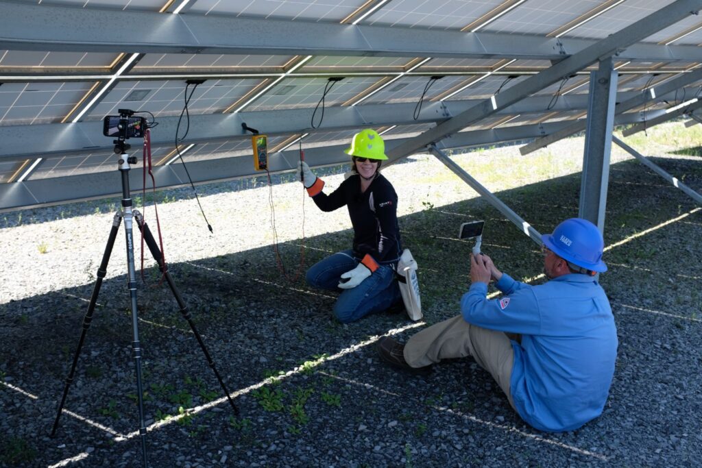 a technician using a multimeter on a solar array and another technician filming
